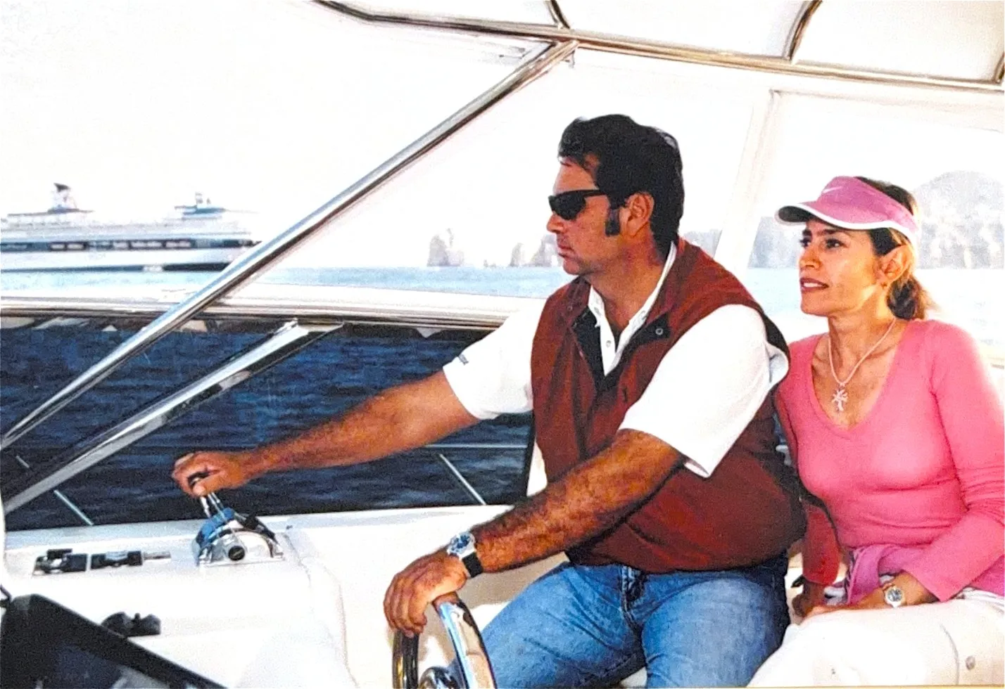 A couple at the helm under the hardtop, cruise ships anchored in the bay behind