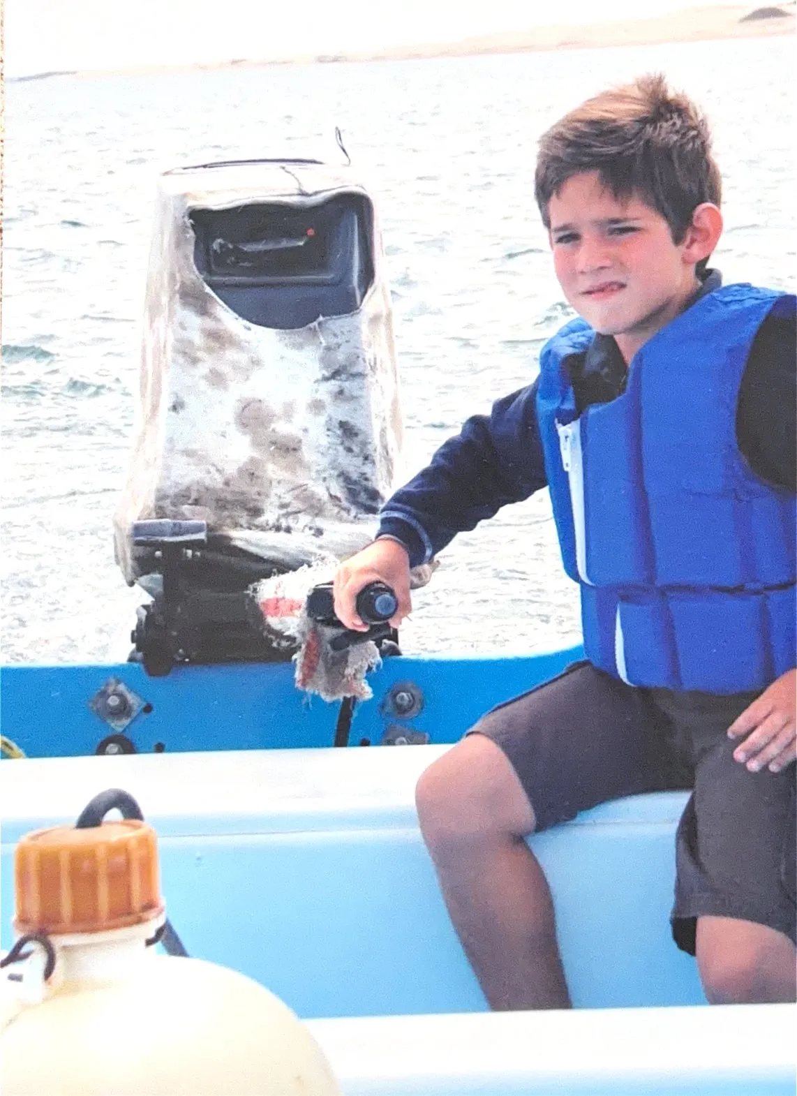 A young Gus Otaduy in a life vest working the tiller of an outboard on a small blue boat