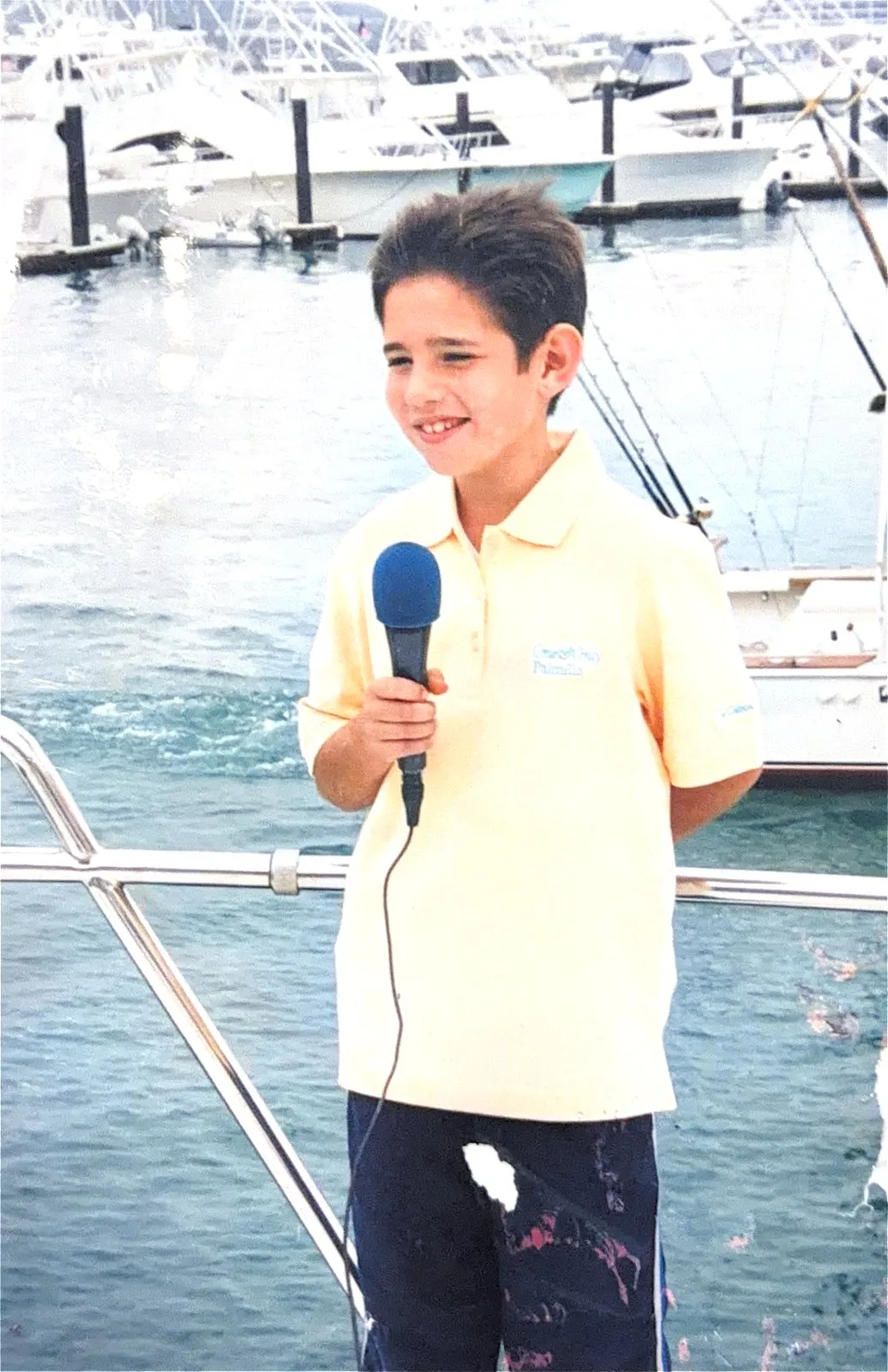 A young Gus Otaduy holding a microphone on a dock at the marina, sportfishers behind