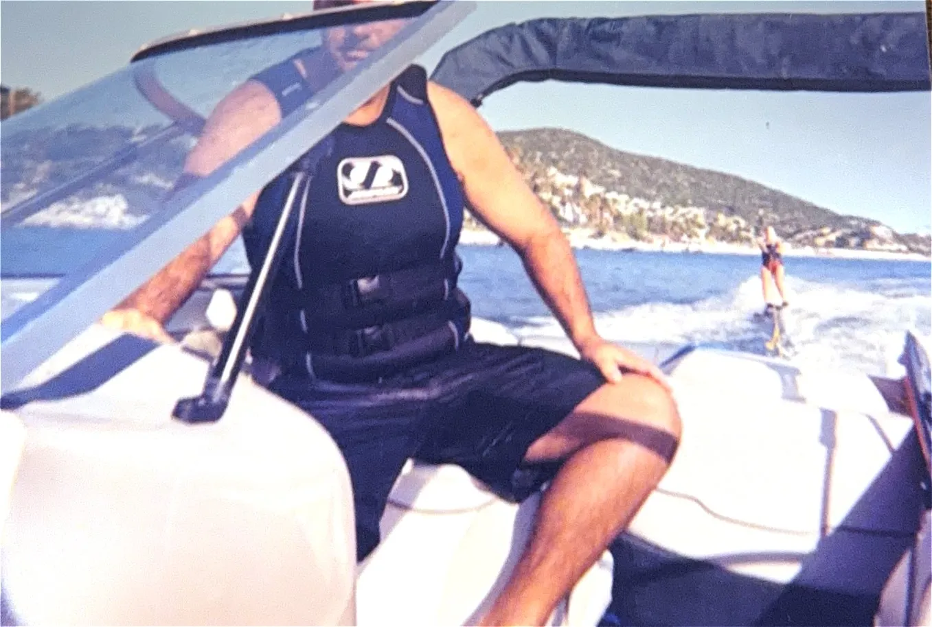 A man in a life vest driving a bowrider, a wakeboarder riding the wake behind