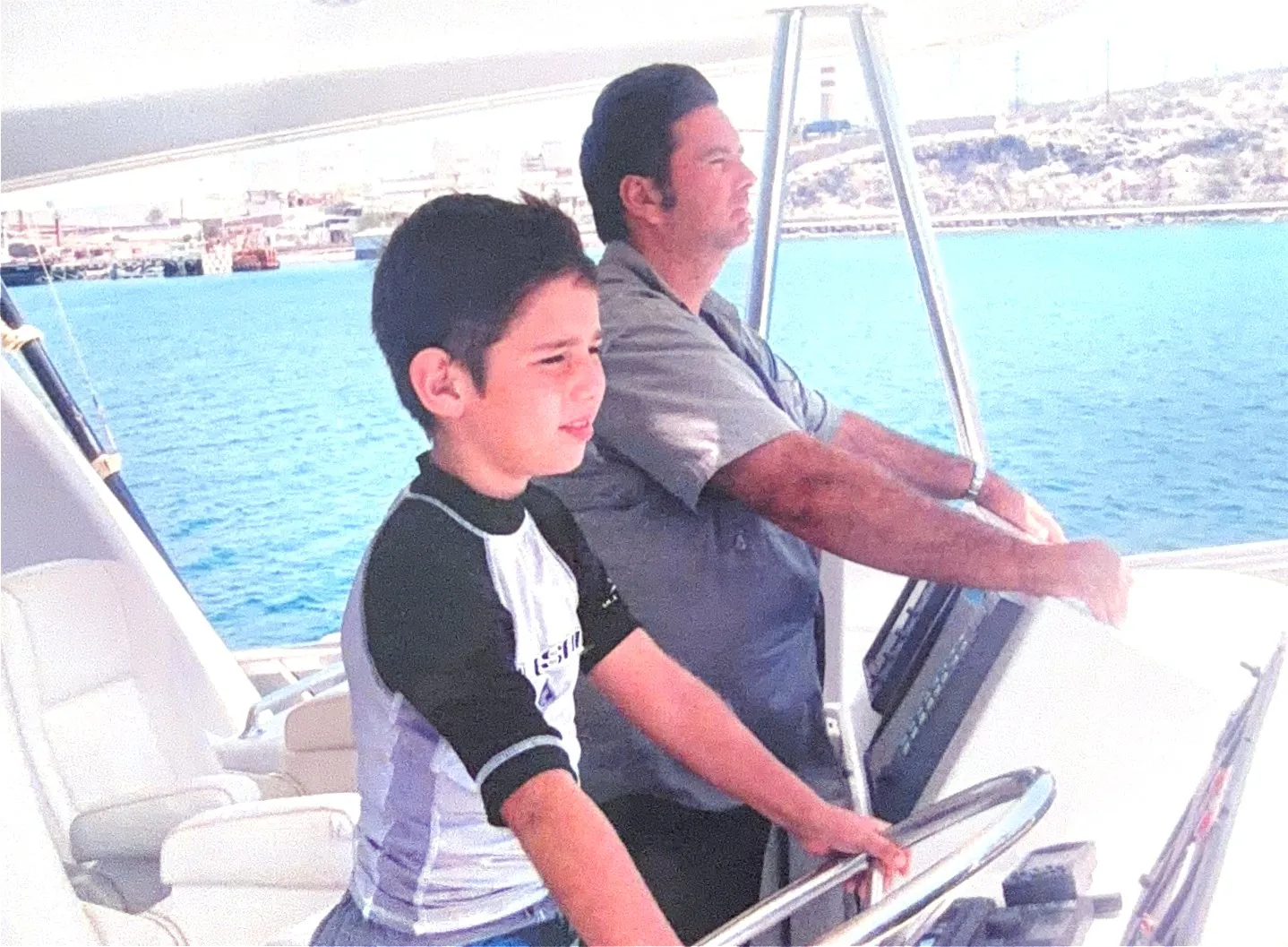 A young Gus Otaduy steering from the flybridge with Augusto Otaduy Sr. standing beside him, the harbor behind
