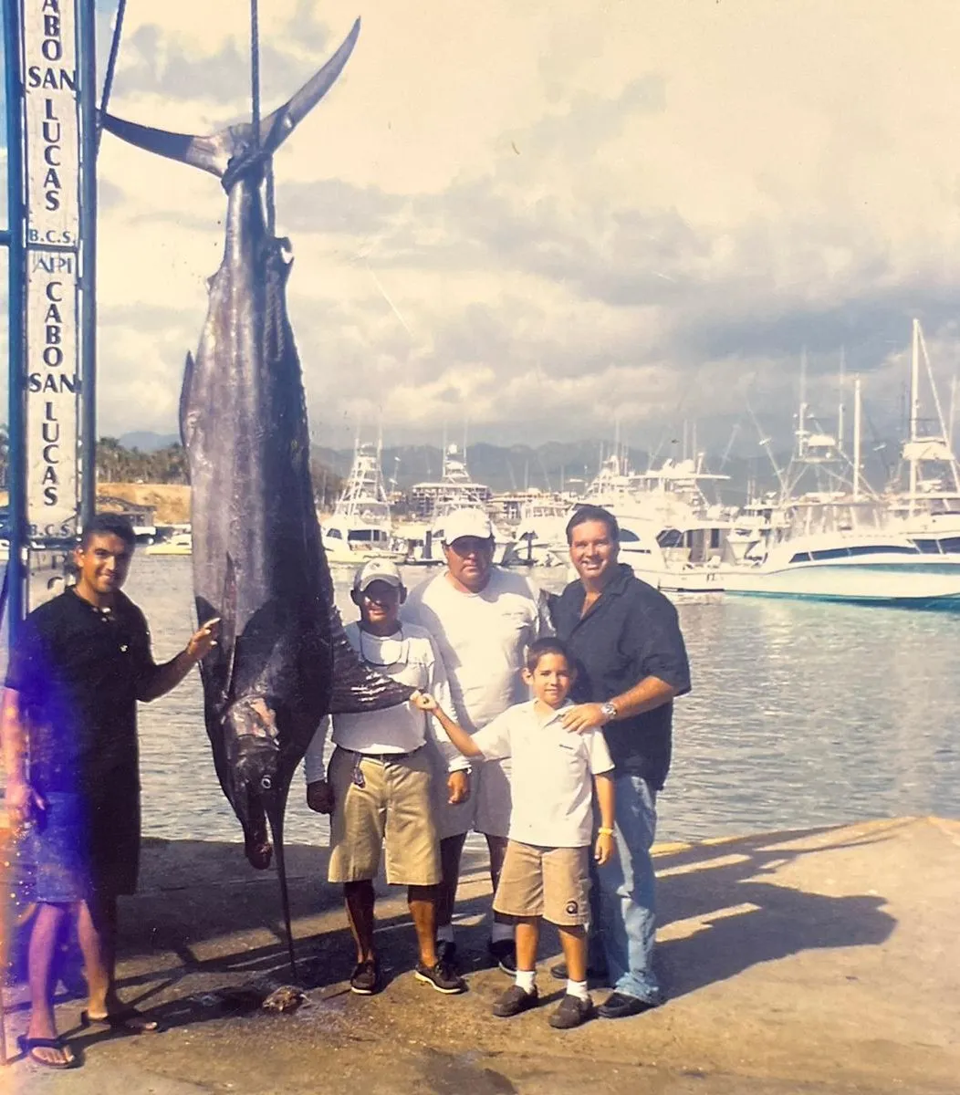 Augusto Otaduy Sr. and his son Gus on the dock at Cabo San Lucas with Captain Tino and Captain Julio, beside a marlin on the scale