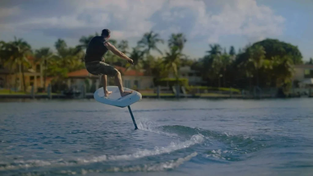 A man in a black shirt and shorts riding a Flite Race Board away leaving streams in the water.